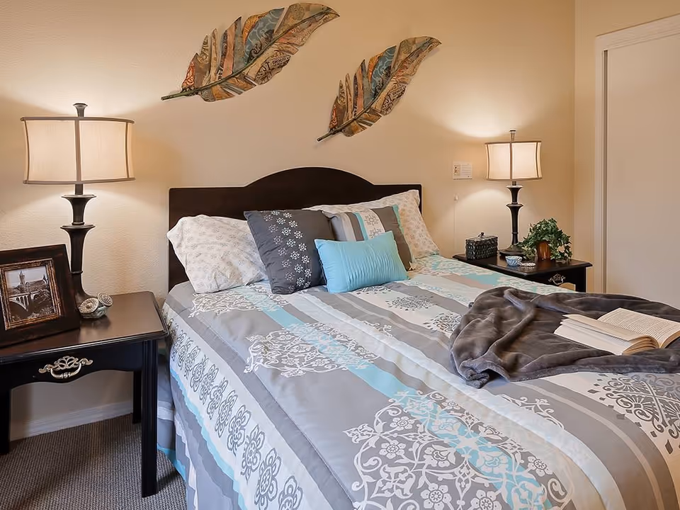 A neatly made bed with a patterned bedspread in shades of gray, white, and light blue, adorned with multiple pillows. On either side of the bed are black nightstands with table lamps, a framed photo, decorative items, and a small plant. Two decorative leaf wall hangings are mounted above the headboard on a beige wall.