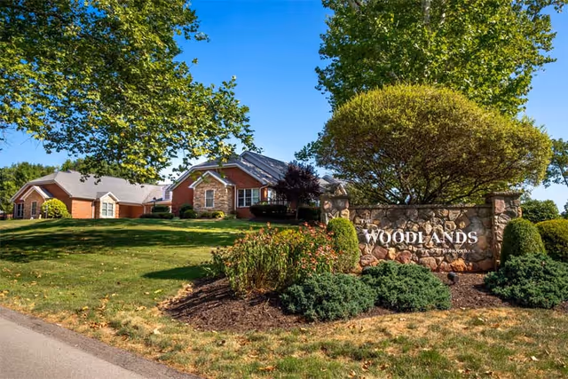 A landscaped outdoor area with a stone sign that reads 'The Woodlands' surrounded by bushes and trees, with residential-style buildings in the background under a clear blue sky.