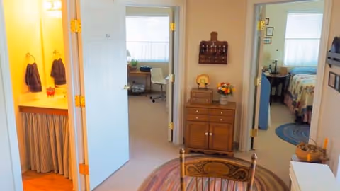 Interior view of a senior living facility hallway with three open doors leading to different rooms. On the left is a bathroom with a sink and towels, straight ahead is a room with a desk and chair, and on the right is a bedroom with a bed and nightstand. A wooden cabinet with decorative items is placed against the wall, and a round rug is on the floor.