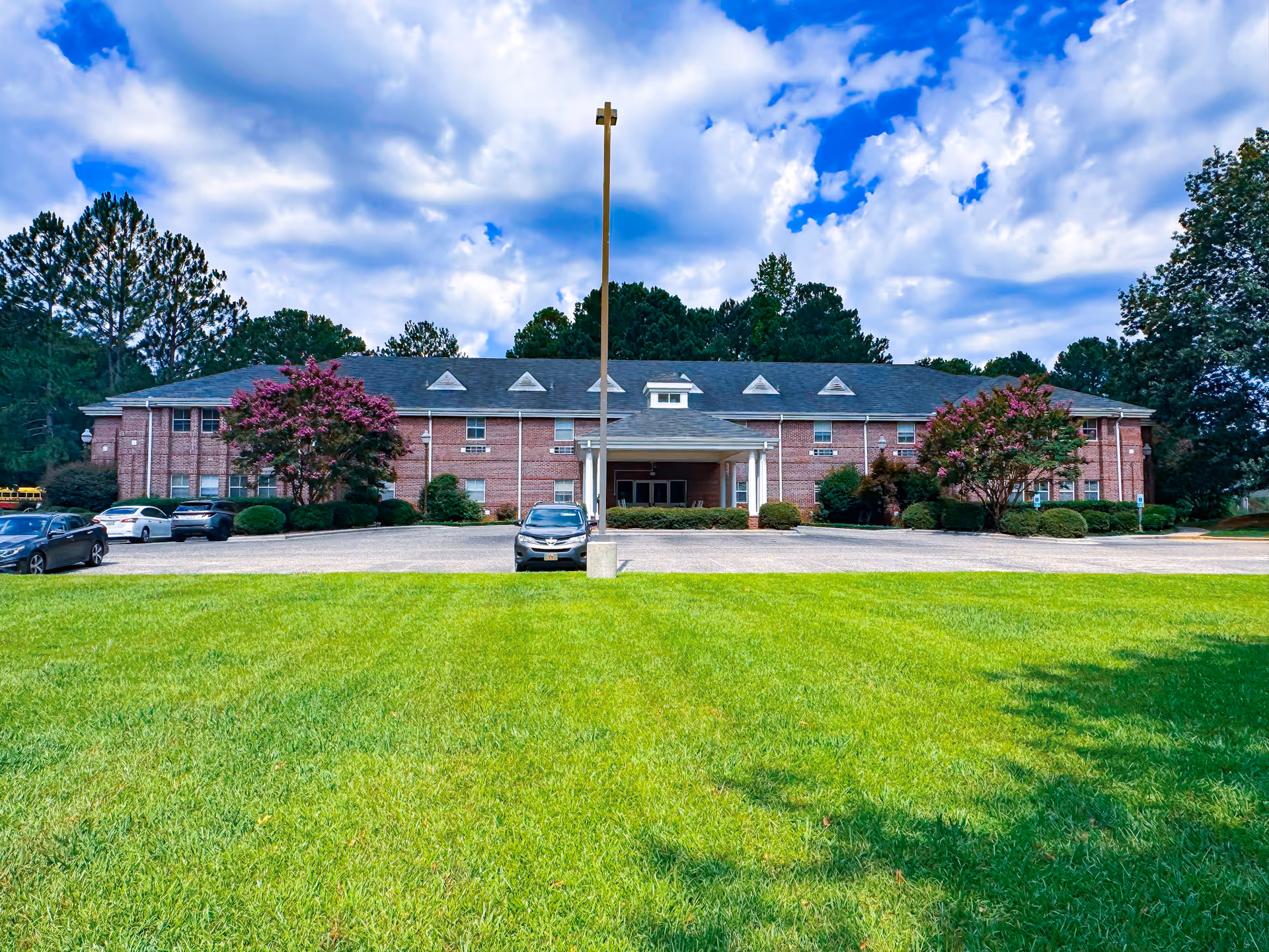Front exterior view of a two-story brick building with a covered entrance, surrounded by green grass, trees, and parked cars under a partly cloudy sky.