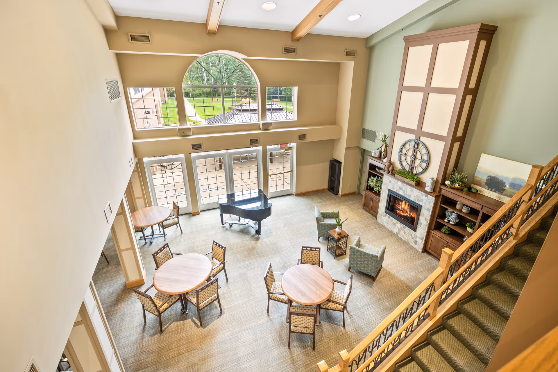 Spacious two-story common lounge with a fireplace, seating areas, round dining tables, and a grand piano viewed from an upper staircase.