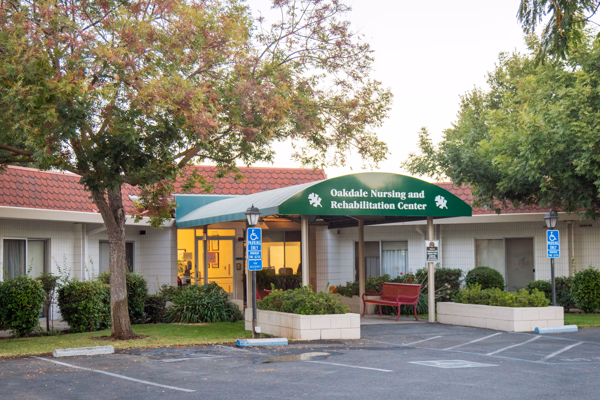 Front entrance of Oakdale Nursing and Rehabilitation Center with a green awning, accessible parking, trees, and a red bench.