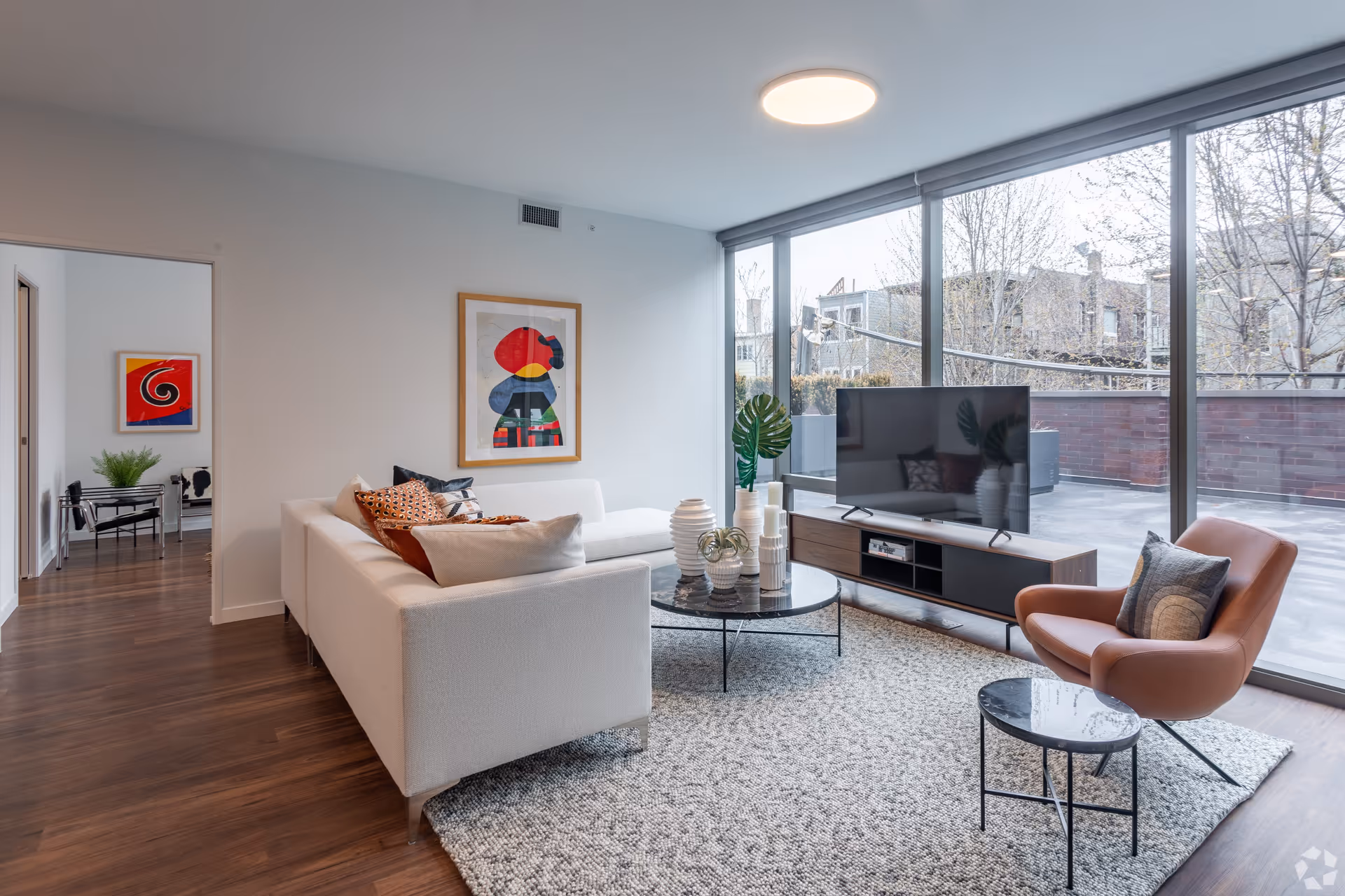 Modern living room with a white sectional sofa adorned with multiple cushions, a round black coffee table with decorative vases, a brown leather armchair with a pillow, and a flat-screen TV on a wooden media console. Large floor-to-ceiling windows provide natural light and a view of an outdoor patio area with trees and buildings in the background. The room has wooden flooring and a textured area rug.
