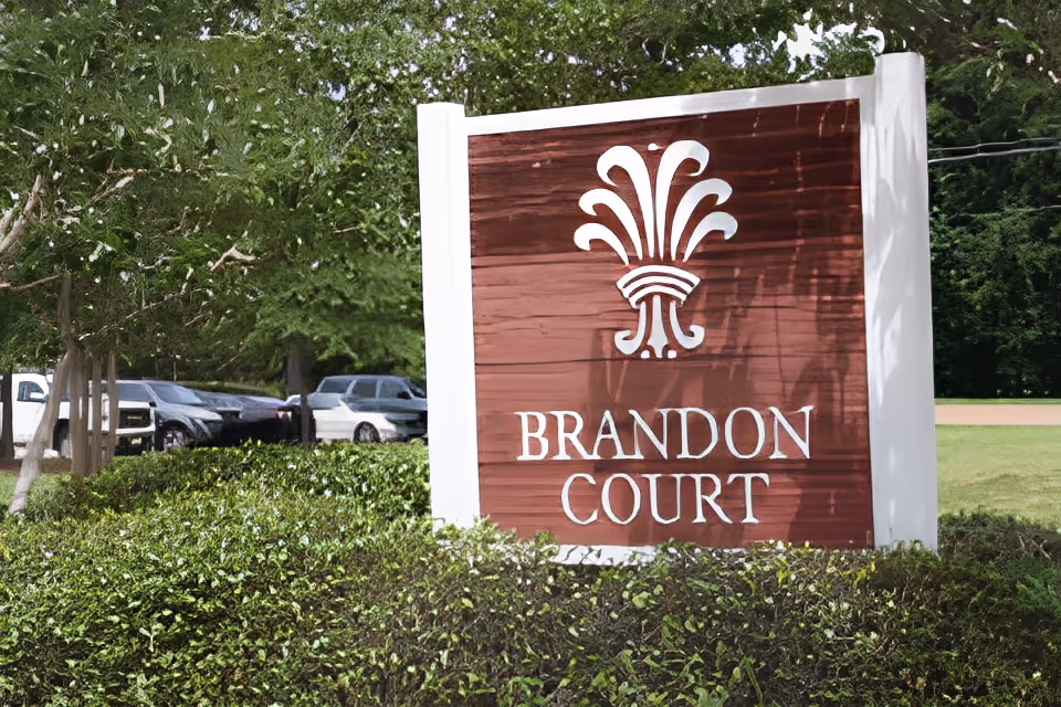 A large wooden sign with white trim displaying the name 'Brandon Court' in white letters, surrounded by green bushes and trees, with parked cars visible in the background.