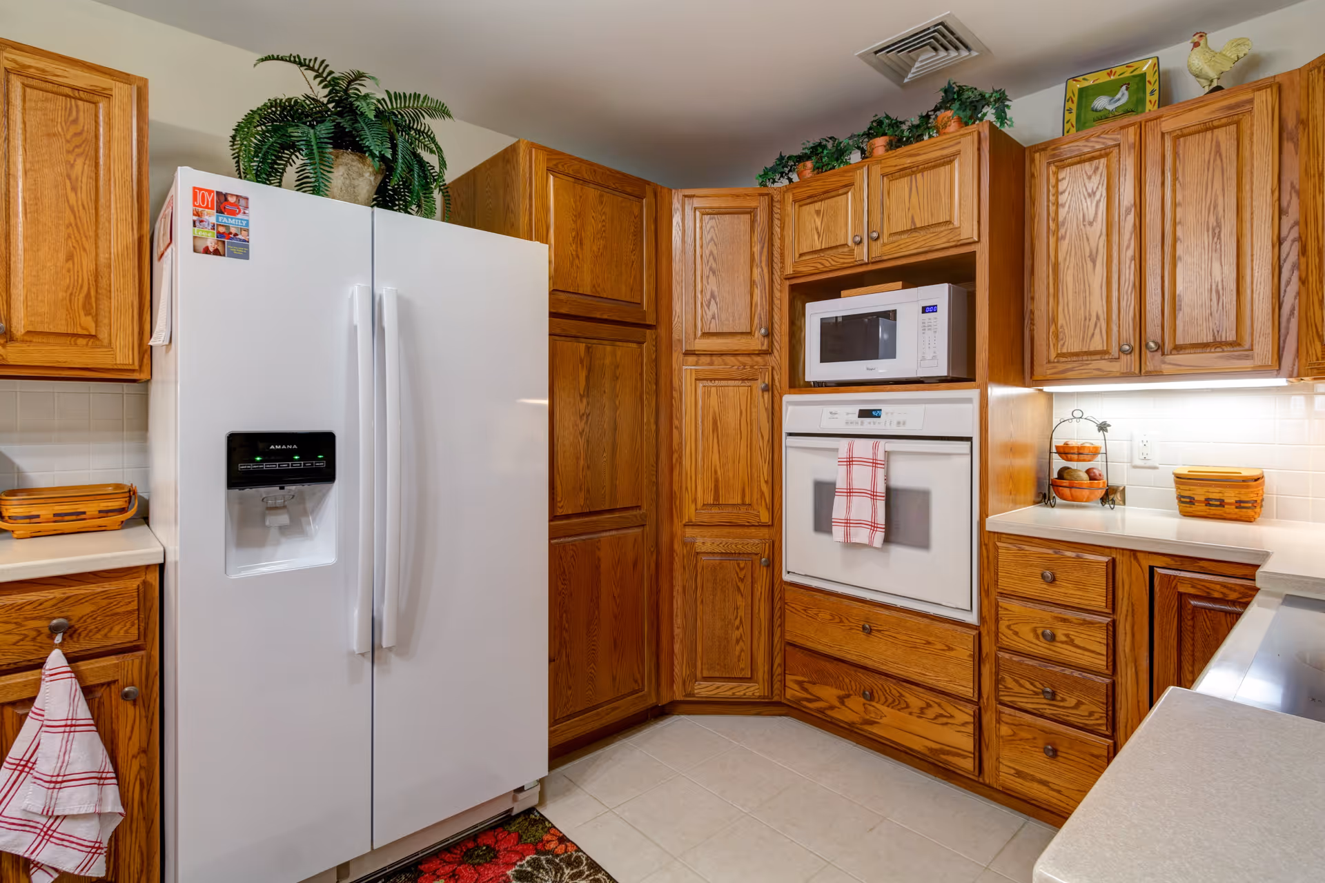 A kitchen with wooden cabinets and white appliances including a side-by-side refrigerator with water dispenser, a built-in microwave, and an oven. The countertops are light-colored, and there are decorative plants and kitchen items on top of the cabinets and counters. A red and white checkered towel hangs on the oven handle.