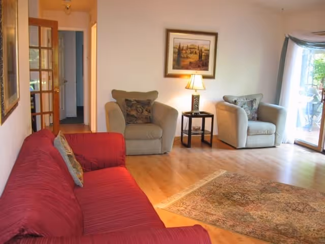 A cozy living room with a red sofa on the left, two beige armchairs with decorative pillows, a small side table with a lamp, a framed landscape painting on the wall, a patterned area rug on the wooden floor, and a sliding glass door leading to an outdoor patio.