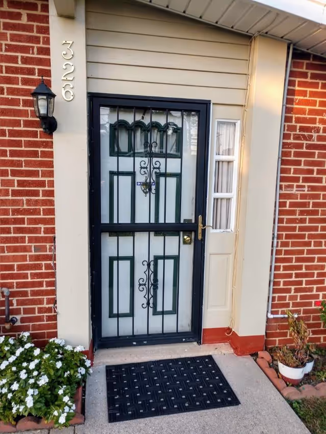 Entrance of a brick house showing a black metal security door over a white decorative door, a wall lantern, vertical house numbers, potted plants, and a black doormat.