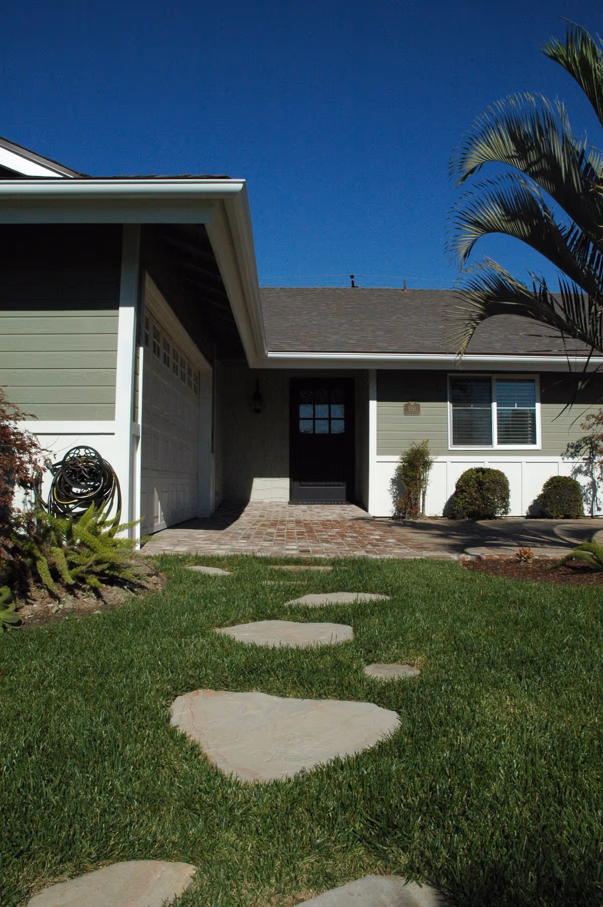 Front entrance of a single-story house with stepping stones across a green lawn leading to a dark front door and attached garage.