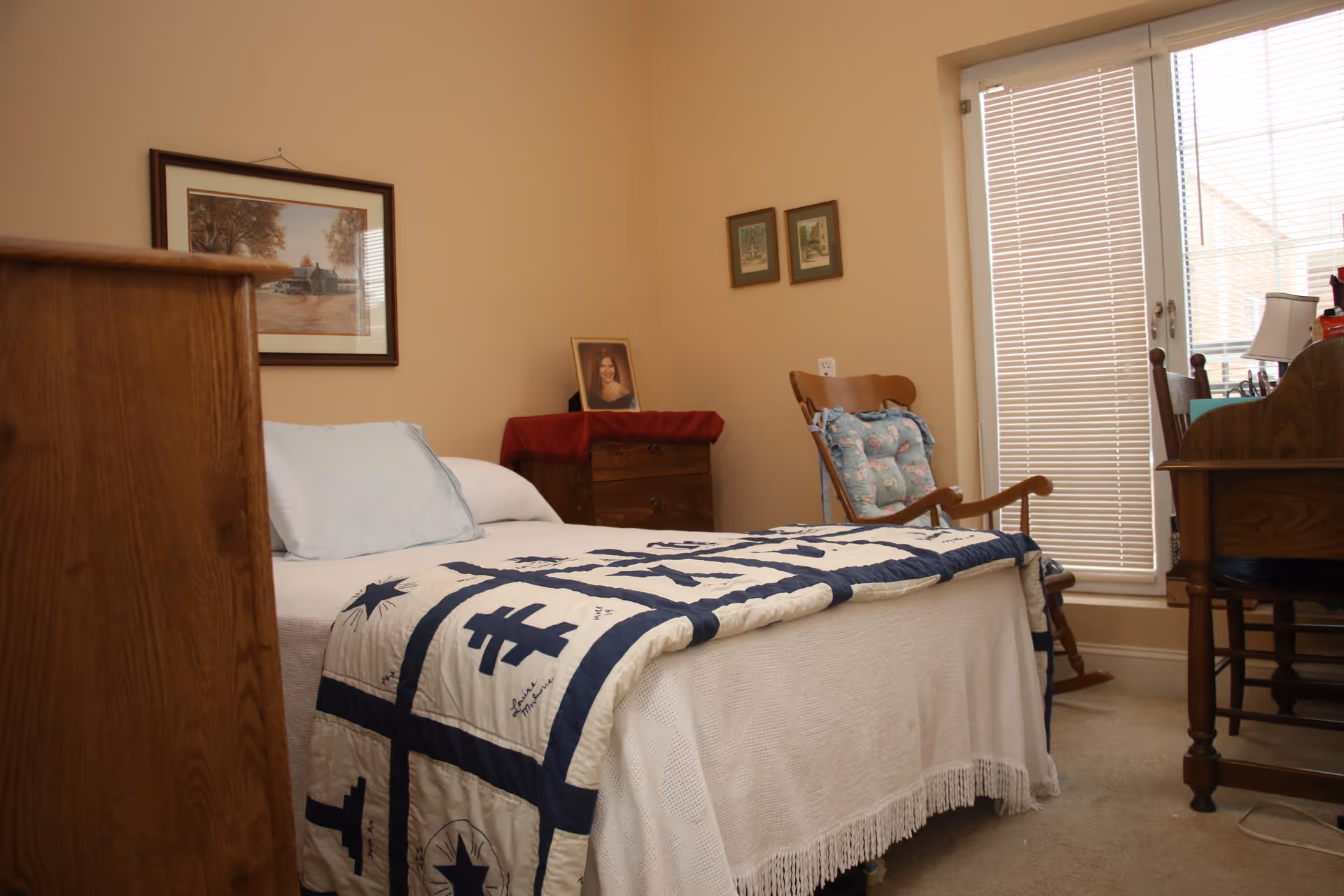 A cozy bedroom with a made bed topped by a patterned quilt, a rocking chair, dresser and a desk by a window with blinds.