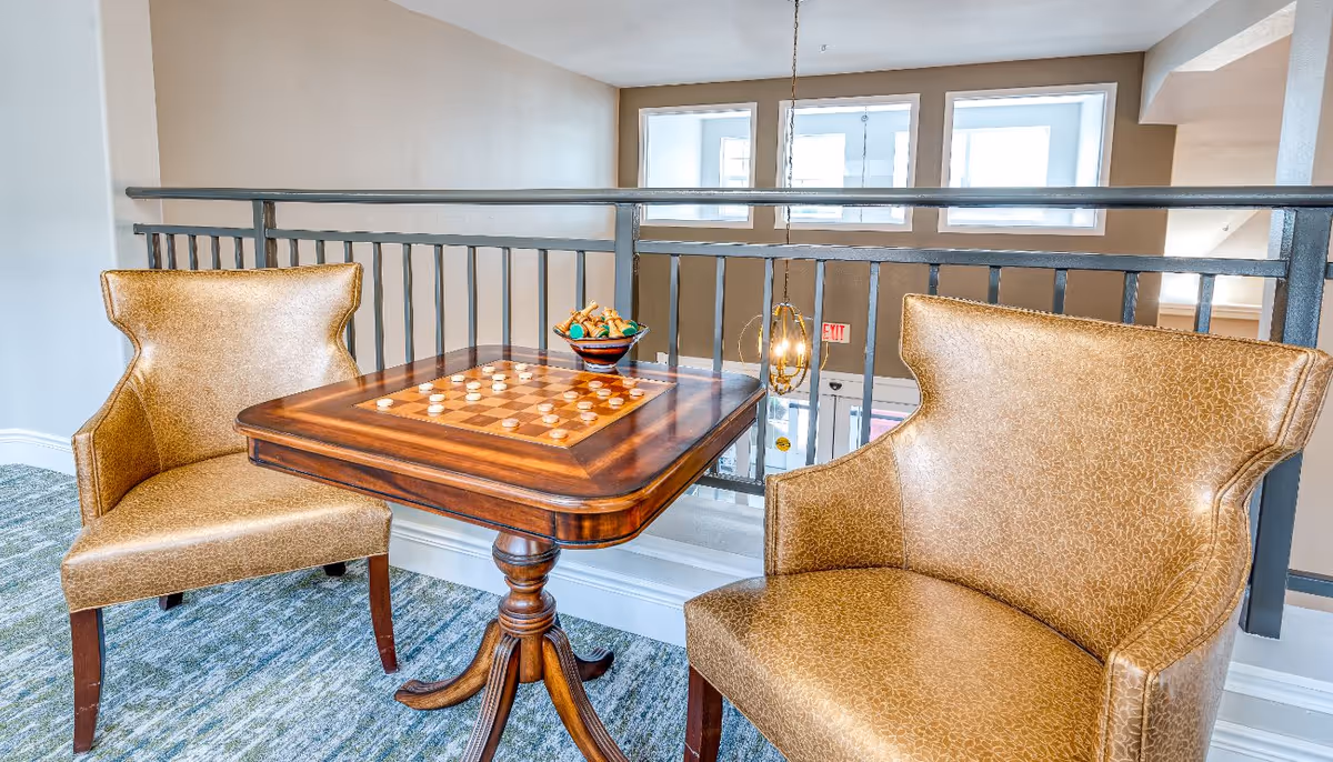 A cozy seating area with two gold-colored armchairs and a wooden table with a built-in checkerboard. A bowl of snacks is placed on the table. The area overlooks a lower level through a black metal railing, with large windows and a hanging light fixture visible in the background.