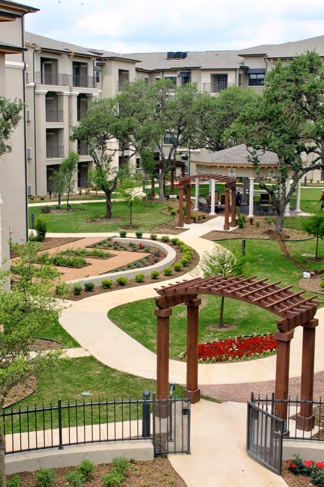 Outdoor courtyard area of a senior living facility with walking paths, green lawns, trees, flower beds, wooden pergolas, and a gazebo surrounded by multi-story residential buildings.