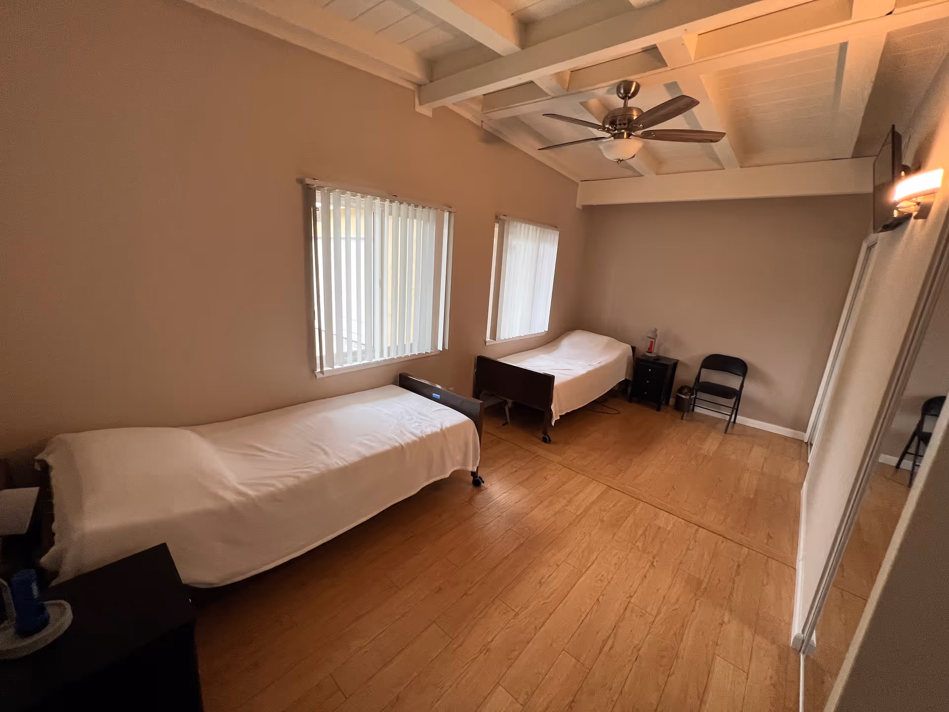 A simple room with two single beds covered in white sheets, each positioned under separate windows with vertical blinds. The room has wooden flooring, a ceiling fan with lights, a small black nightstand between the beds, a black folding chair, a wall-mounted TV, and a large mirror leaning against the wall.