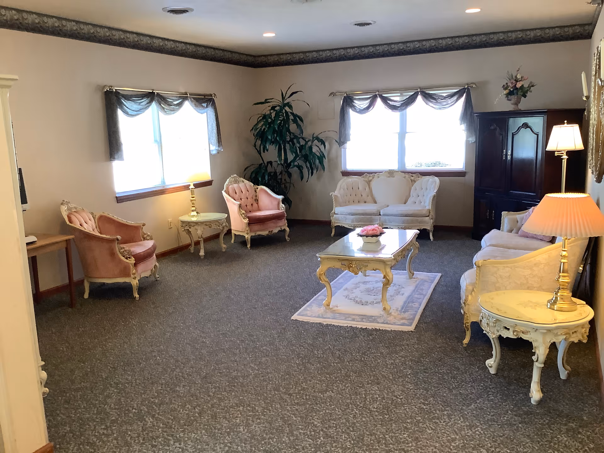 Sunlit common living room with vintage pink and cream upholstered chairs and sofas arranged around a central coffee table.