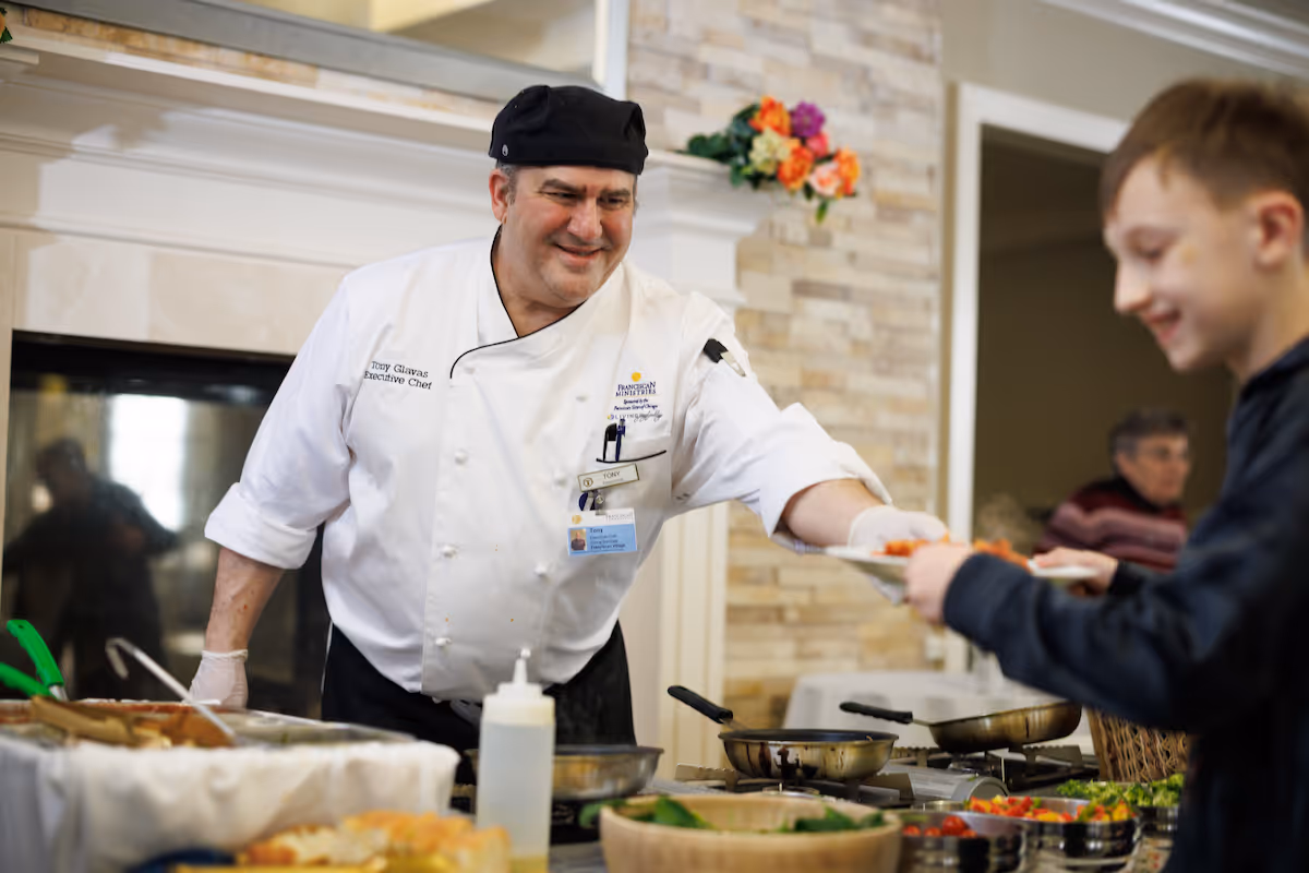 A chef wearing a white uniform and black hat is serving food to a smiling young boy at a buffet-style setup inside a dining area with a stone wall and a fireplace in the background.