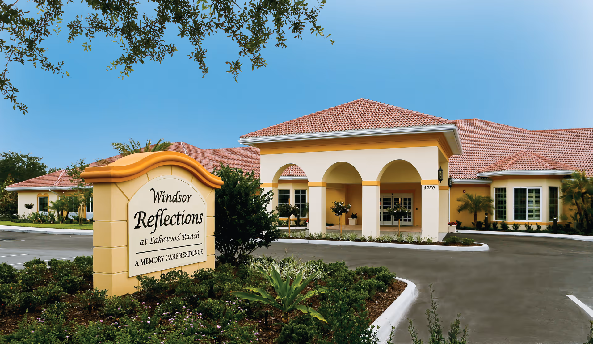 Exterior front view of Windsor Reflections Memory Care Of Lakewood Ranch building with a sign in the foreground displaying the facility name. The building has a tiled roof, arched entrance, and surrounding landscaping with plants and trees under a clear blue sky.