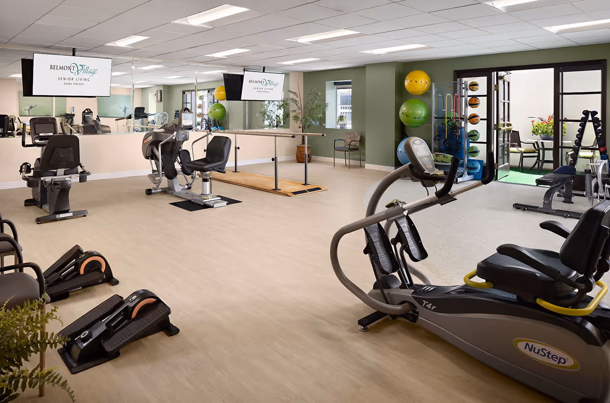A spacious senior living fitness room at Belmont Village Senior Living Sabre Springs featuring various exercise equipment including recumbent bikes, elliptical machines, and step platforms. The room has light wood flooring, green walls, large mirrors, and two flat-screen TVs mounted on the wall displaying the Belmont Village logo. There is also a rack with colorful exercise balls and weights, and a door leading to an outdoor patio area with seating and plants.