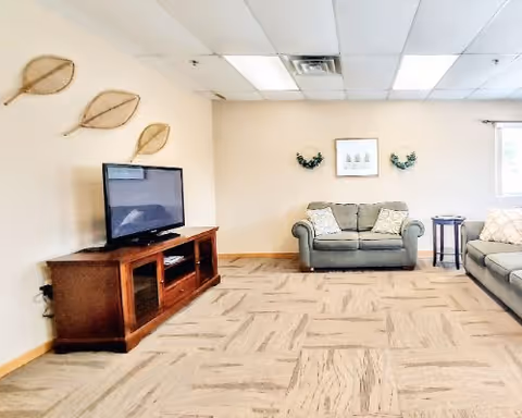 A cozy living room with beige walls and carpeted floor. The room features a flat-screen TV on a wooden TV stand to the left, a gray loveseat with two patterned pillows in the center, and a gray sofa with a white pillow on the right. There are decorative wall hangings including three leaf-shaped pieces above the TV and two small wreaths flanking a framed picture above the loveseat. A small round side table is placed between the loveseat and sofa. The ceiling has fluorescent lighting panels.