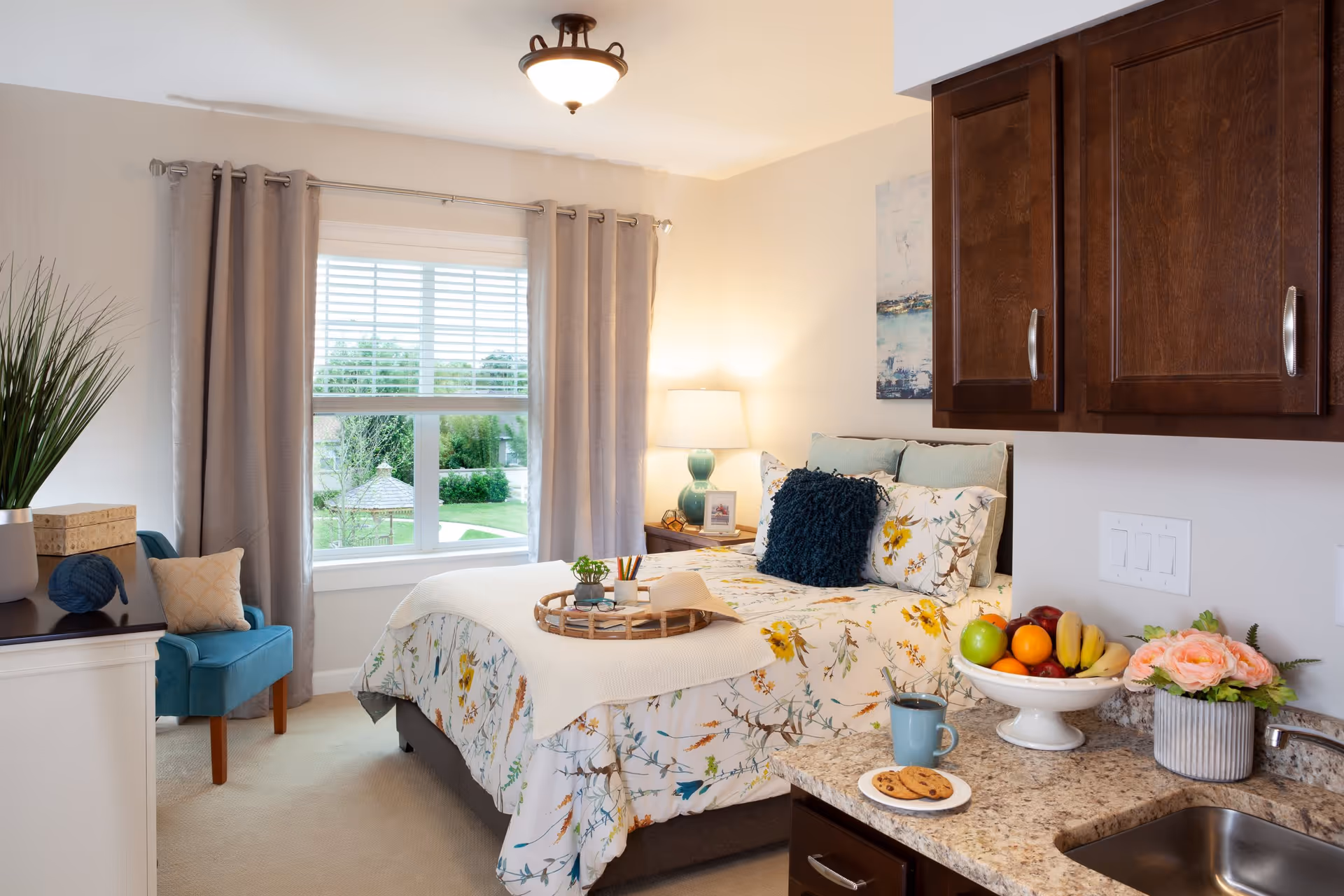 Well-lit bedroom studio with a bed dressed in floral bedding, a window with curtains, a blue chair, and a small kitchenette counter with fruit and flowers.