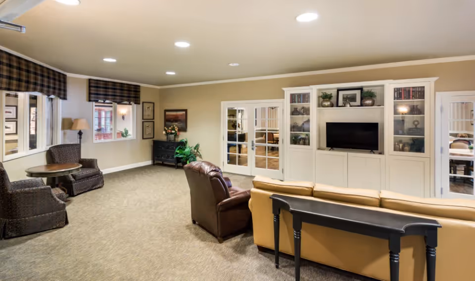 Spacious senior living common room with sofas and chairs facing a built-in TV cabinet and French doors.