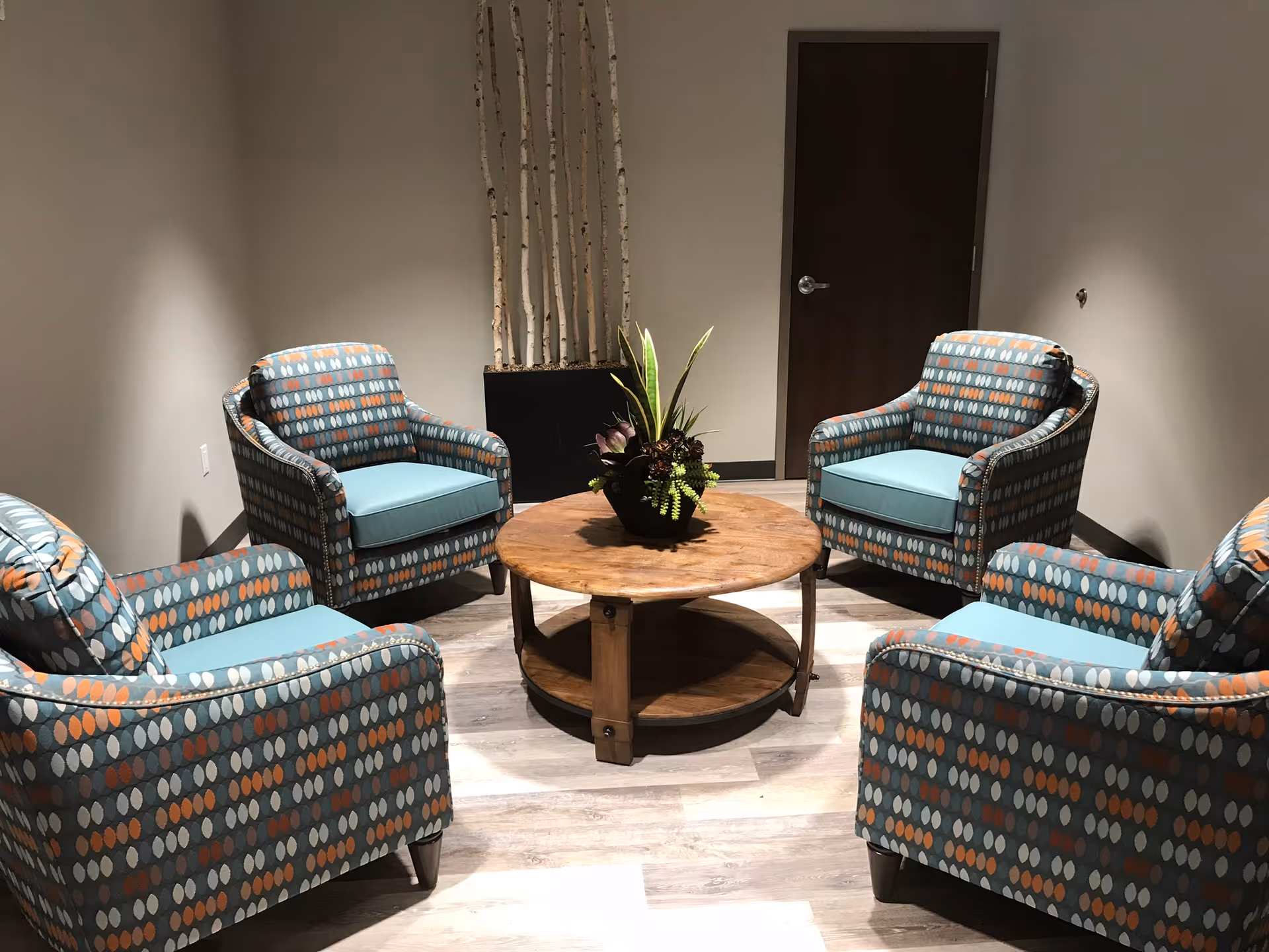 A small seating area with four patterned armchairs arranged around a round wooden coffee table with a plant centerpiece. The room has light-colored walls, a wooden door, and a tall black planter with decorative birch branches in the corner.