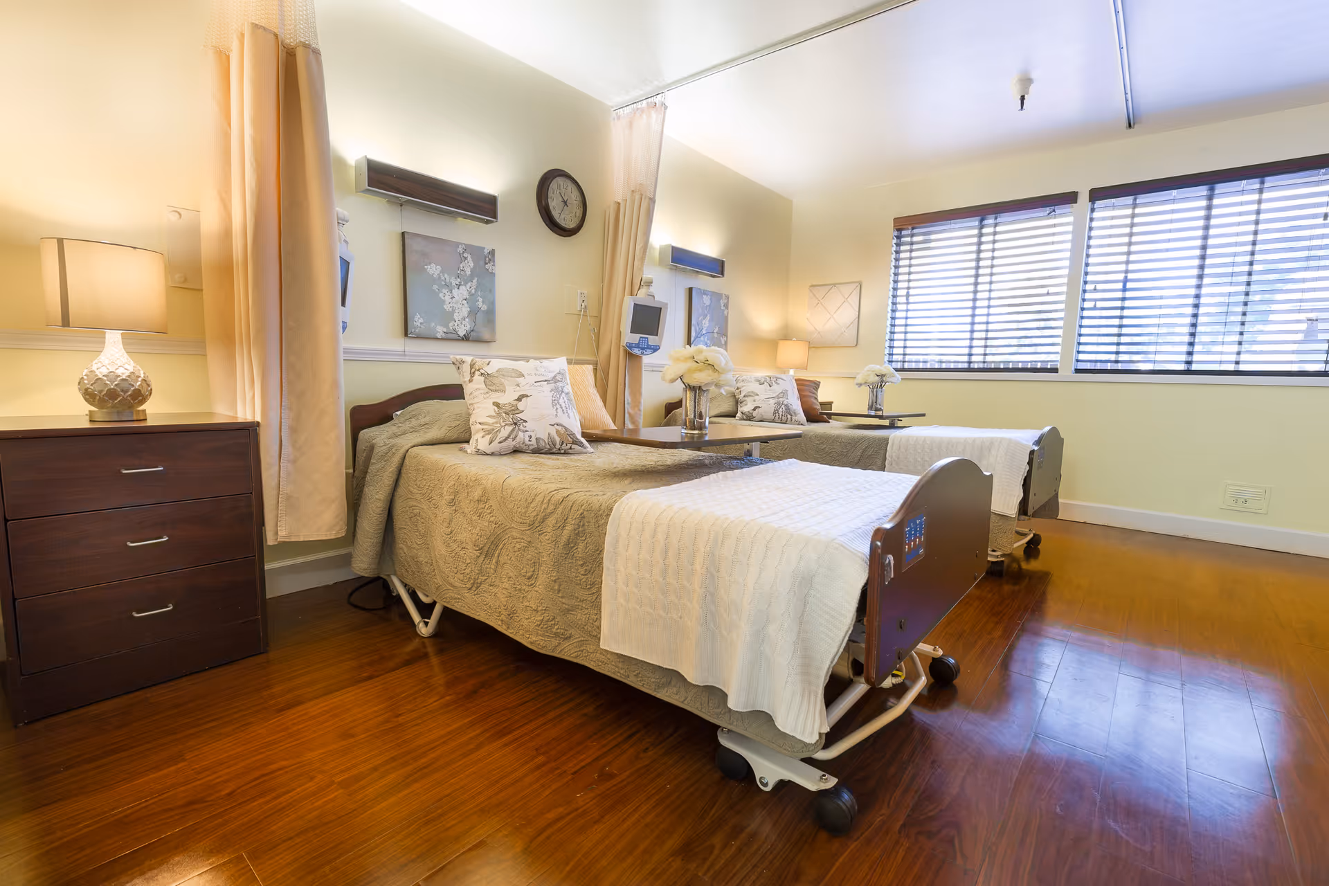 A bright and clean room with two hospital-style beds, each with beige and white bedding, decorative pillows, and overbed tables with flower vases. The room has wooden flooring, a nightstand with a lamp, wall-mounted medical equipment, a clock, and large windows with blinds letting in natural light.