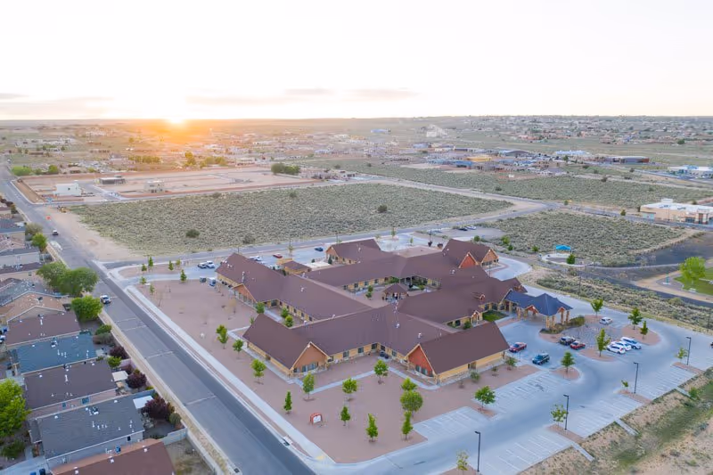 Aerial view of a single-story senior living complex with connected brown-roofed buildings, parking areas, and surrounding desert landscape at sunset.