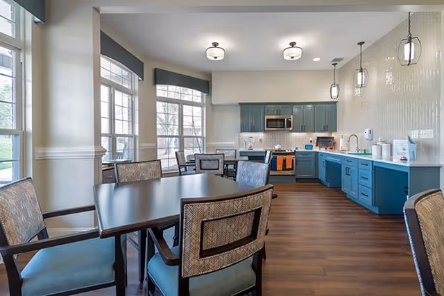 A bright and modern kitchen and dining area with large windows letting in natural light. The kitchen features blue cabinetry, a white countertop, stainless steel appliances including a microwave and stove, and pendant lighting. The dining area has a dark wooden table surrounded by cushioned chairs with patterned backs. The floor is a warm wood tone.
