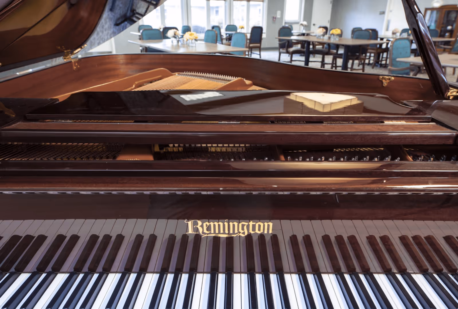 Close-up view of a Remington grand piano keyboard and strings with a dining area featuring tables and chairs visible in the background.