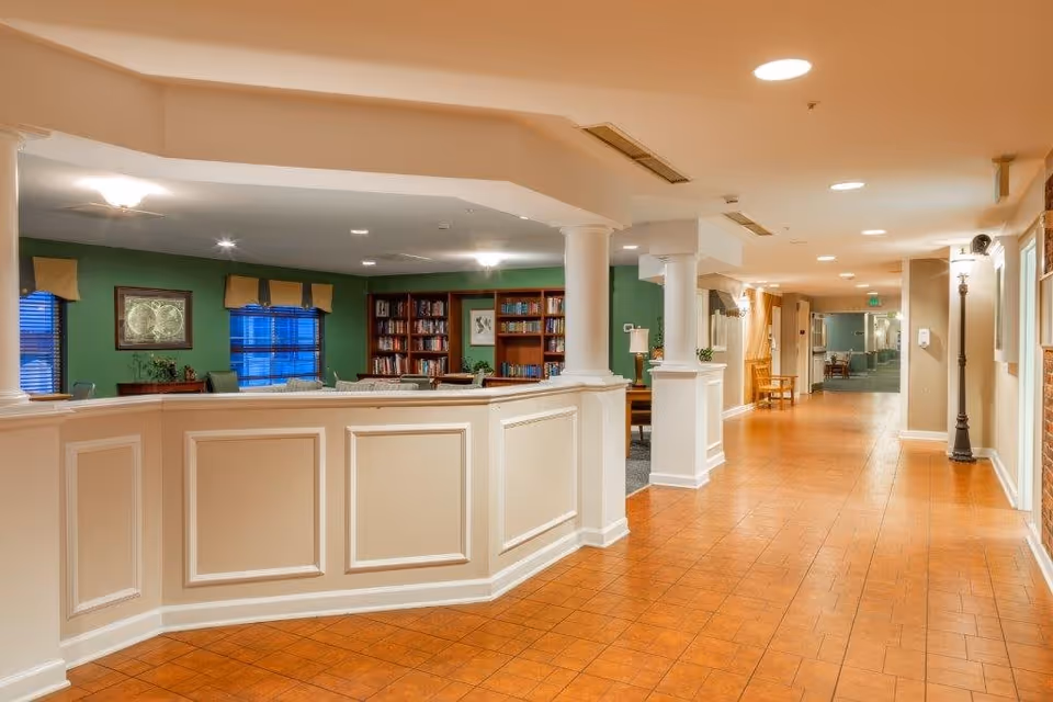 Interior view of a senior living facility hallway with a partial white paneled wall and columns on the left side, leading into a room with green walls, bookshelves filled with books, and windows with beige valances. The hallway has a tiled floor and ceiling lights, with a bench and lamp visible further down the corridor.