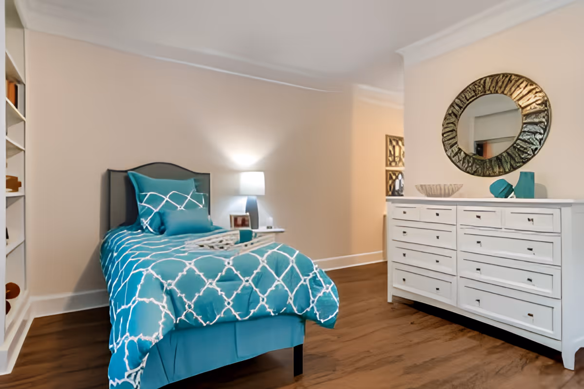A tidy bedroom with a single bed dressed in teal patterned bedding, a bedside lamp, and a white dresser topped by a round mirror.