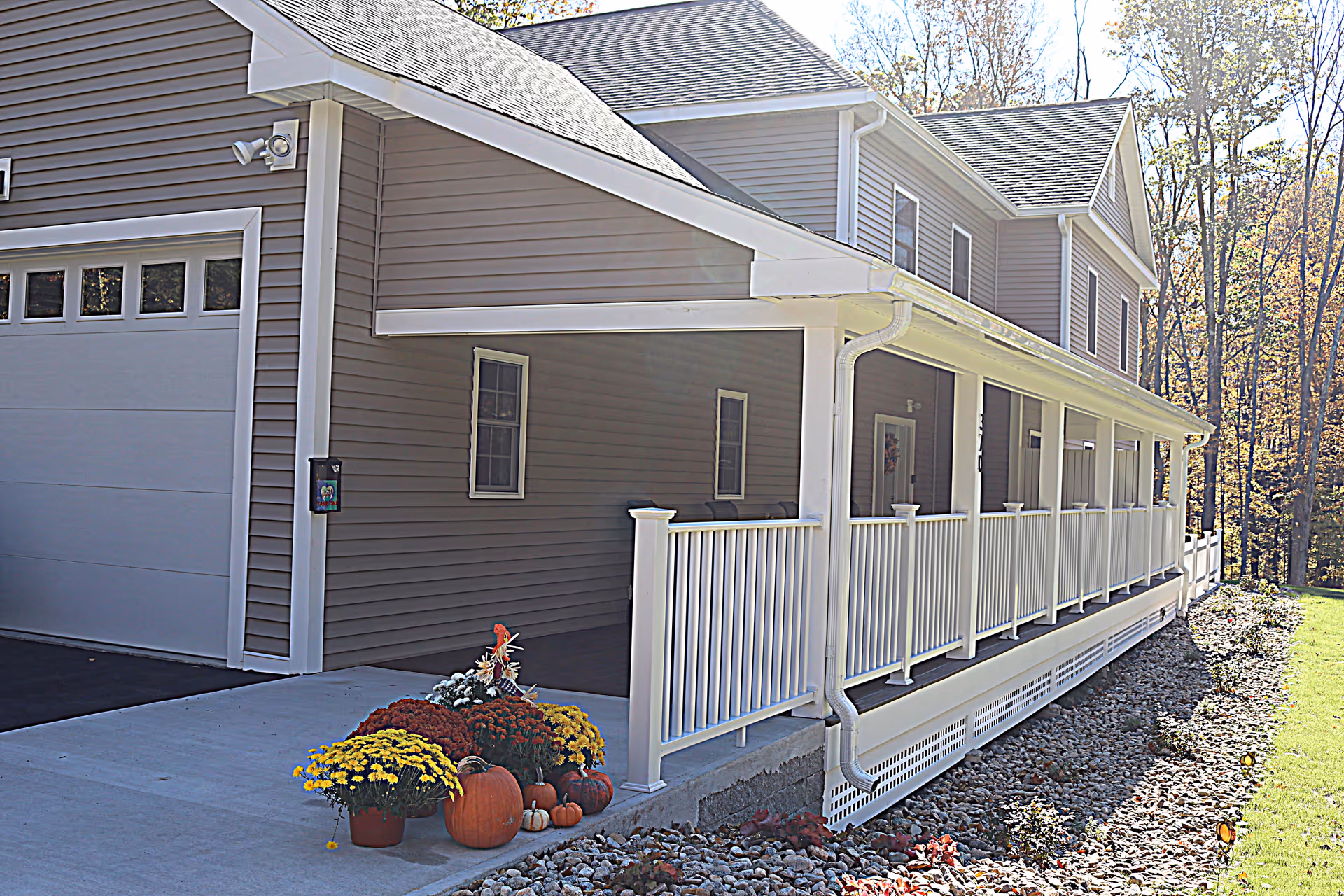 Exterior view of a residential care home with gray siding and a white railing along a covered porch. There are autumn decorations including pumpkins and potted flowers near the driveway. Trees and grass are visible in the background.
