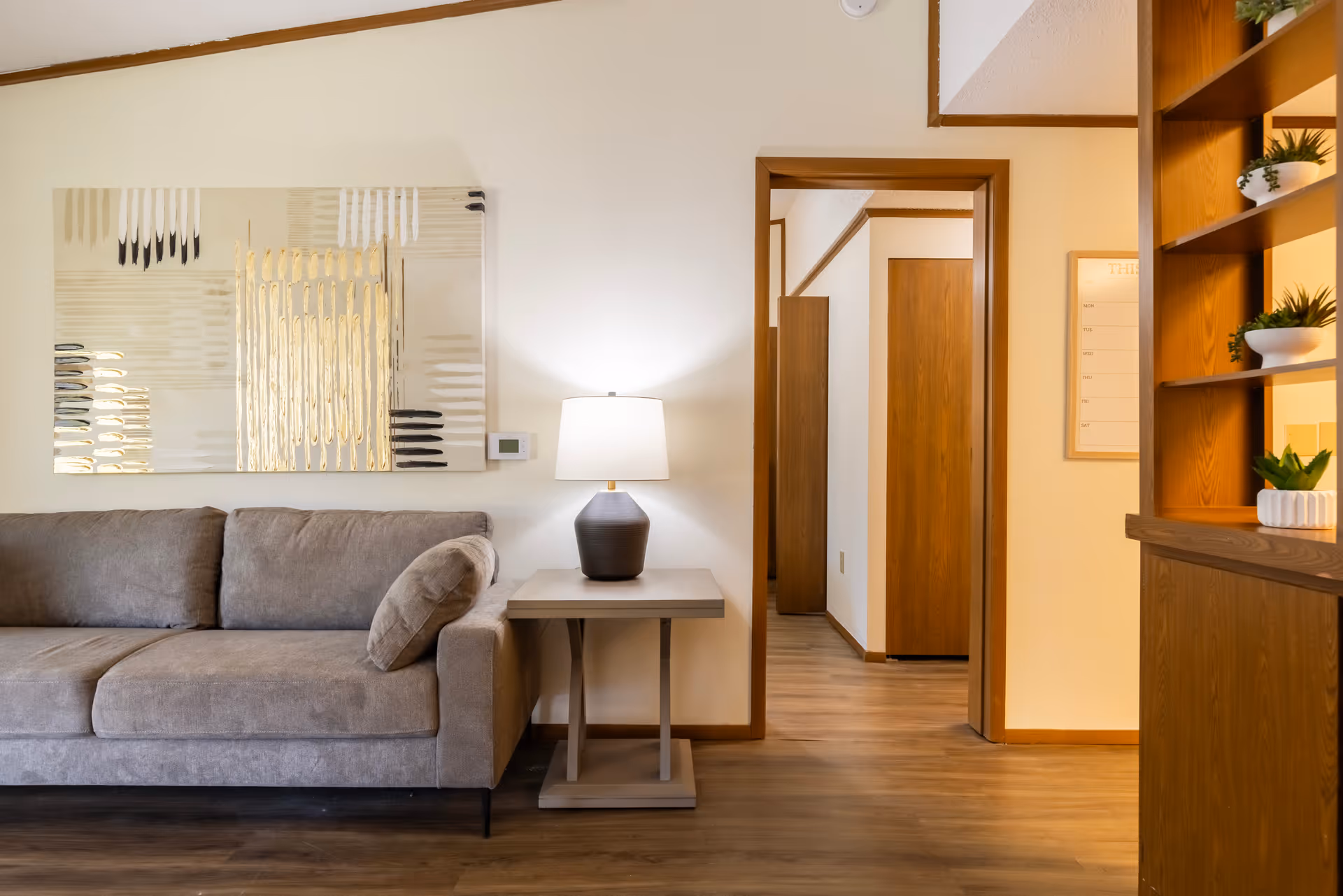 A cozy living room area with a gray sofa and a matching pillow, a side table with a lamp, a large abstract painting on the wall, and a wooden floor. There is an open doorway leading to a hallway with wooden doors and a wooden shelving unit with potted plants on the right.