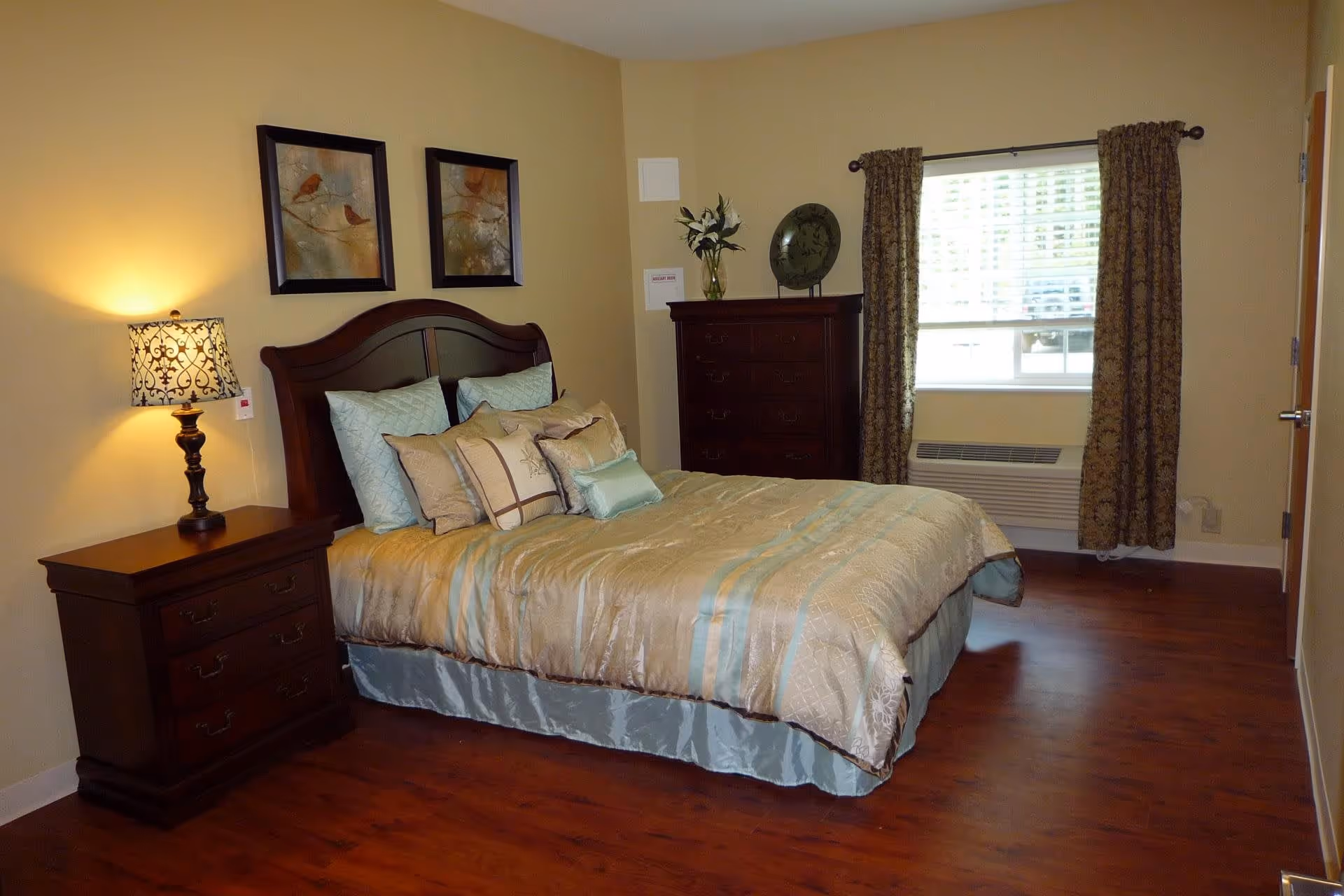 A neatly made bed with multiple pillows in a bedroom featuring wooden furniture including a nightstand with a decorative lamp and a tall dresser. The room has beige walls, a window with patterned curtains, and wooden flooring.