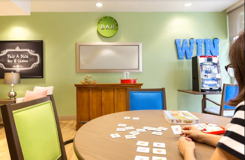 A person playing a memory card game at a round table in a brightly lit room with green walls. The room has colorful chairs, a wooden cabinet, a slot machine, and wall decorations including a green clock labeled 'BAR' and blue letters spelling 'WIN'. A framed sign on the wall reads 'Pair A Dice Bar & Casino'.