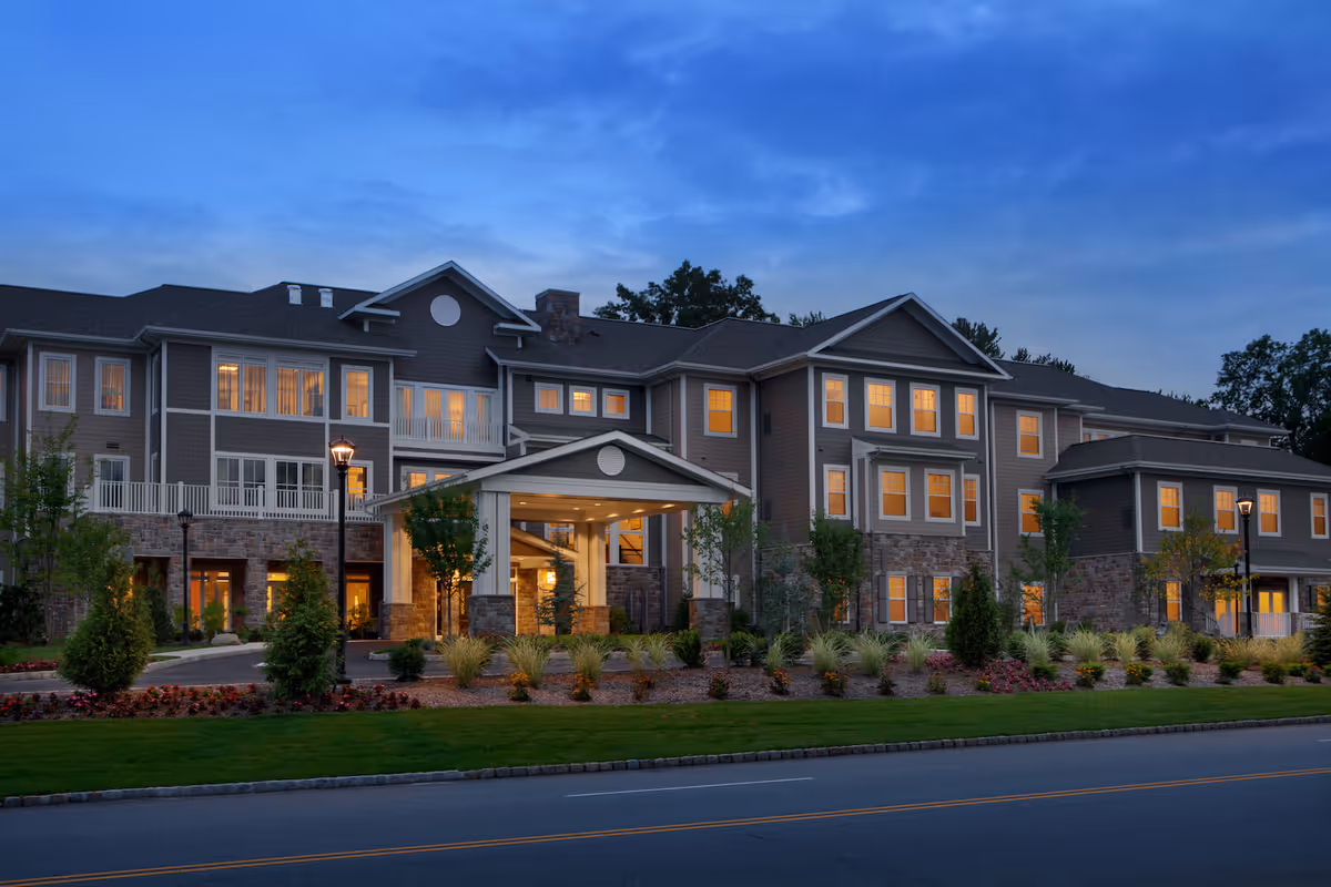 Exterior view of a large, multi-story senior living facility building at dusk with warm lights glowing from the windows. The building features a covered entrance with stone pillars, landscaped gardens, and a paved road in front.