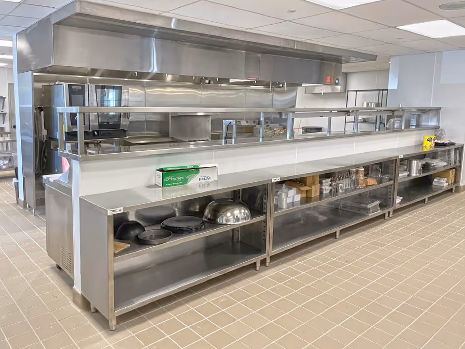 A clean, modern commercial kitchen with stainless steel counters and shelves. Various kitchen utensils, plates, and containers are neatly arranged on the shelves. The kitchen features a large exhaust hood and cooking equipment in the background.