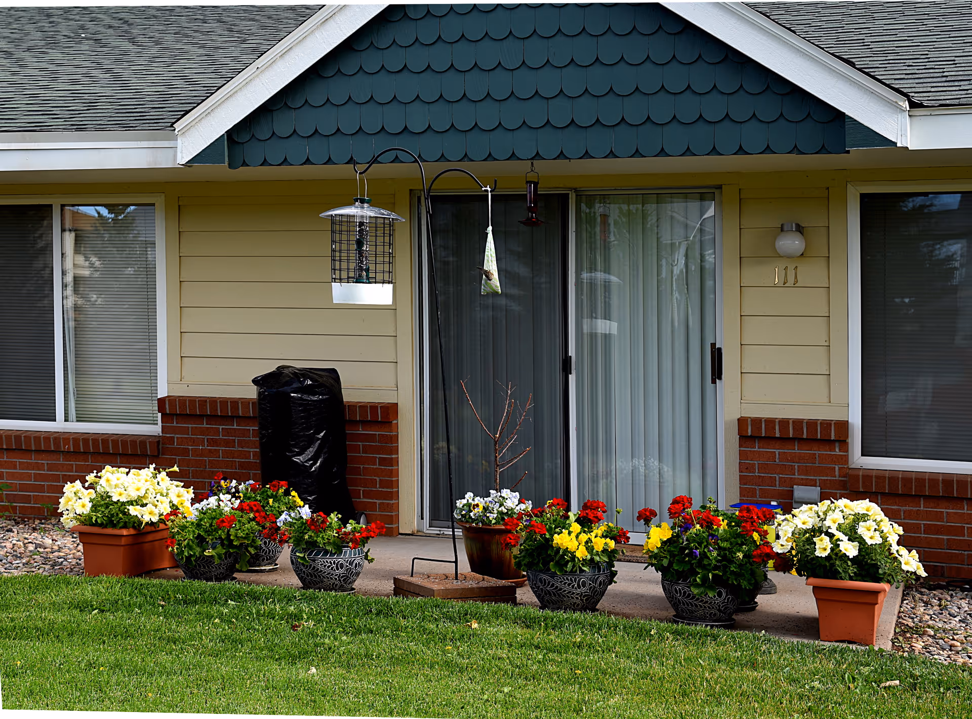 A ground-level front patio with a sliding glass door, hanging bird feeders and several pots of colorful flowers on the lawn.