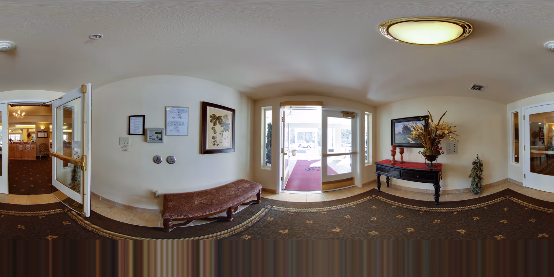 Interior view of the entrance area in Oak Park Senior Living by Cogir, featuring a glass door leading outside, a cushioned bench against the wall, framed artwork, a decorative table with a floral arrangement, and carpeted flooring with tile accents.