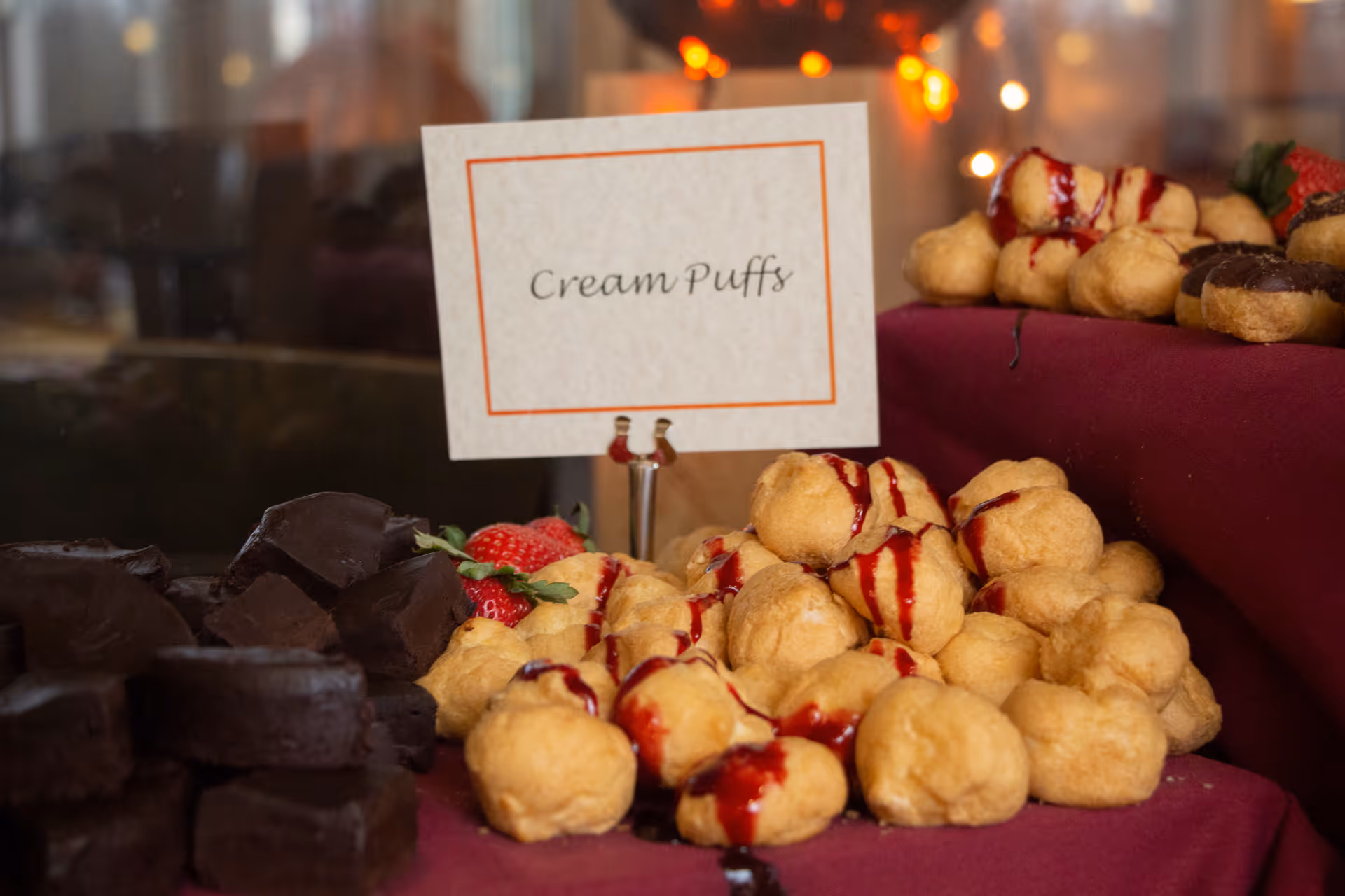 A platter of cream puffs drizzled with sauce next to a sign reading "Cream Puffs" and other desserts on a table.