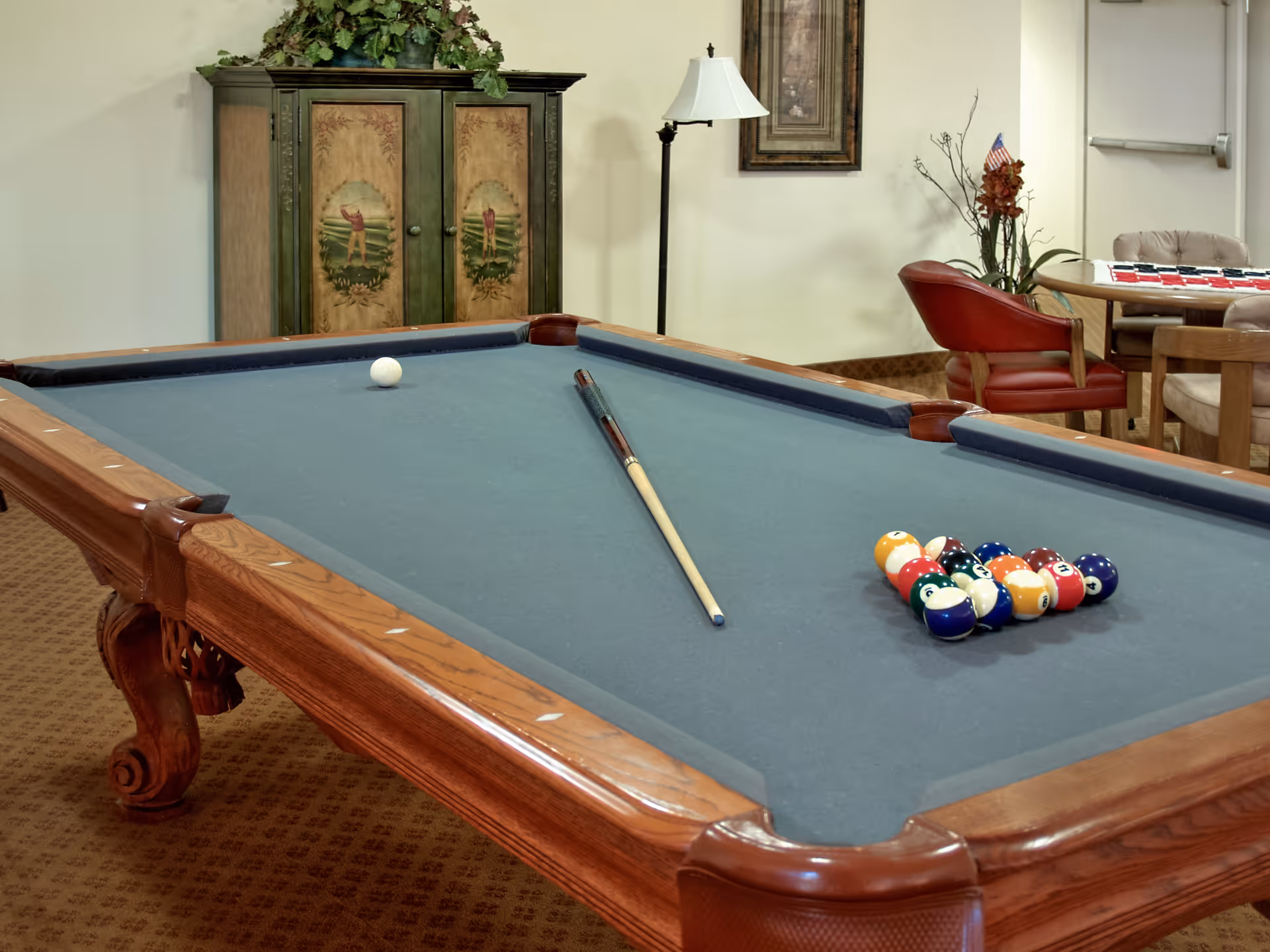 A billiards room with a pool table featuring a blue felt surface, a cue stick, a white cue ball, and a rack of colorful billiard balls. In the background, there is a decorative cabinet with painted panels, a floor lamp, a framed picture on the wall, and a seating area with red and beige chairs around a small table.