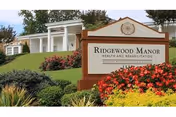 Entrance sign for Ridgewood Manor Health and Rehabilitation surrounded by well-maintained landscaping with colorful flowers and greenery, with a white building featuring columns visible in the background.