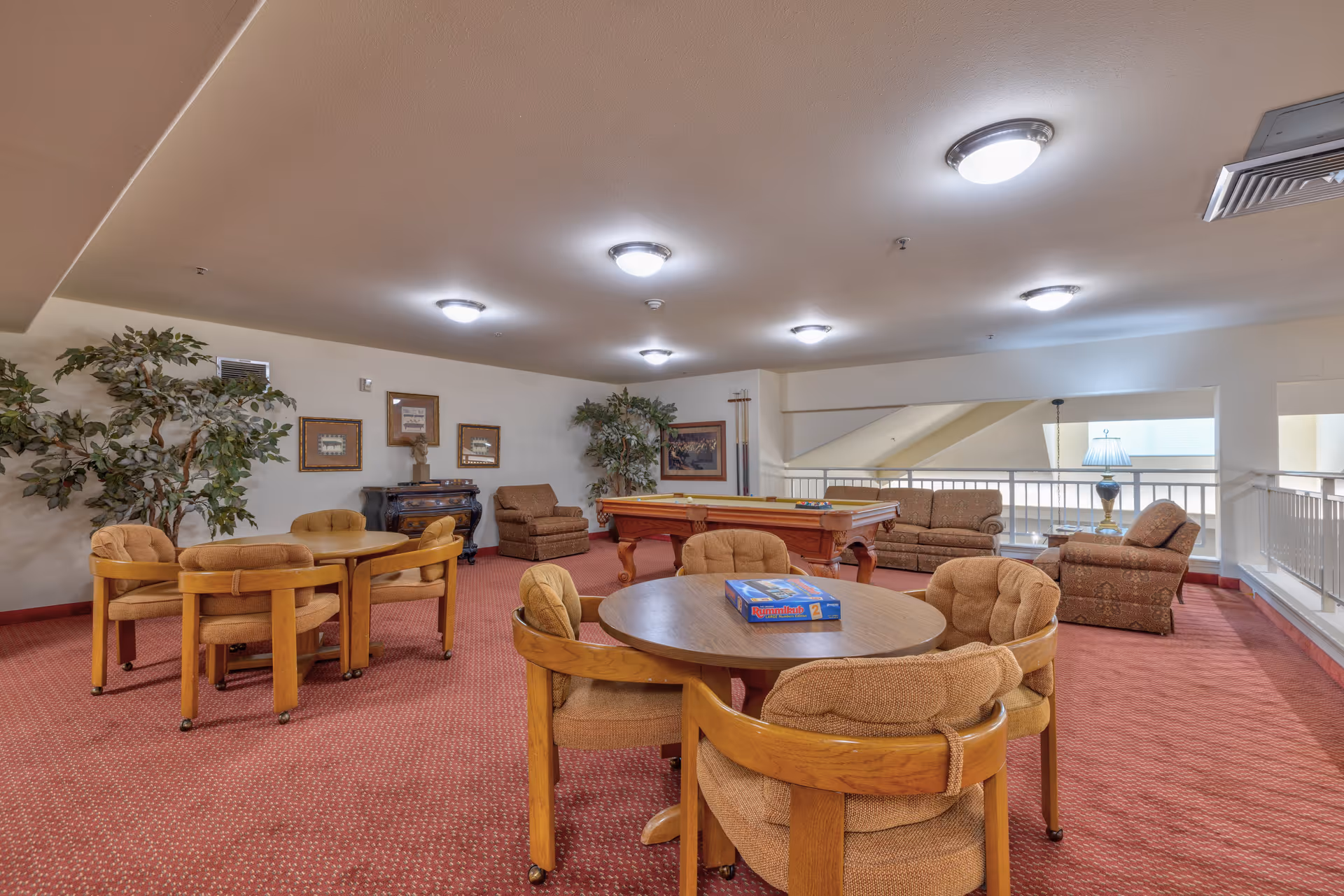 A cozy common area with round wooden tables and cushioned chairs on a red carpeted floor. There is a pool table in the background, along with several armchairs and a sofa. The walls are decorated with framed pictures and there are large potted plants adding greenery to the space. The ceiling has multiple round light fixtures providing bright illumination.