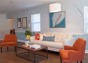 A bright and modern living room with a white sofa adorned with colorful pillows, two orange armchairs, a wooden coffee table with books and decorative items, a potted plant, and framed artwork on the light blue walls. Large windows allow natural light to fill the space.