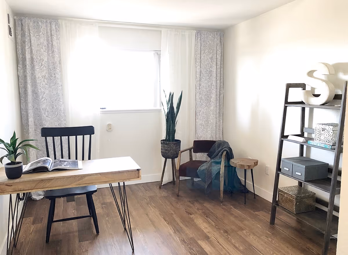 Bright furnished living room/office with a wooden desk and chair, potted plants, a lounge chair with a throw, side table and shelving against white walls.