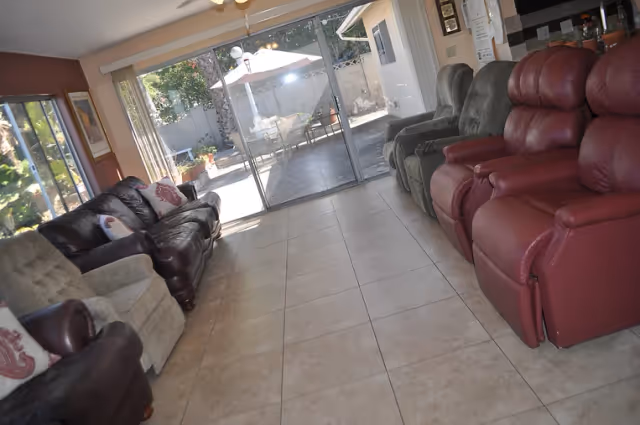 A living room area with several recliner chairs and a sofa arranged along the walls. The room has tiled flooring and large sliding glass doors that open to an outdoor patio with a table, chairs, and an umbrella. The space is well-lit with natural light coming through the windows and glass doors.
