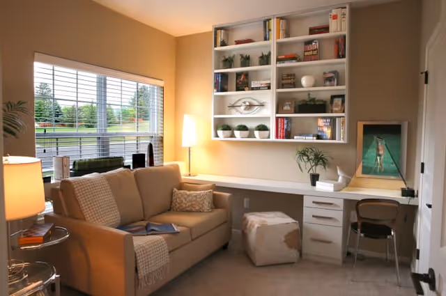 A cozy living room area with a beige sofa adorned with pillows and a throw blanket, a floor lamp beside it, and a large window with blinds letting in natural light. On the opposite wall, there is a built-in white desk with drawers, a chair, a small ottoman, and a wall-mounted bookshelf filled with books and decorative items. A framed picture and a small plant are on the desk.