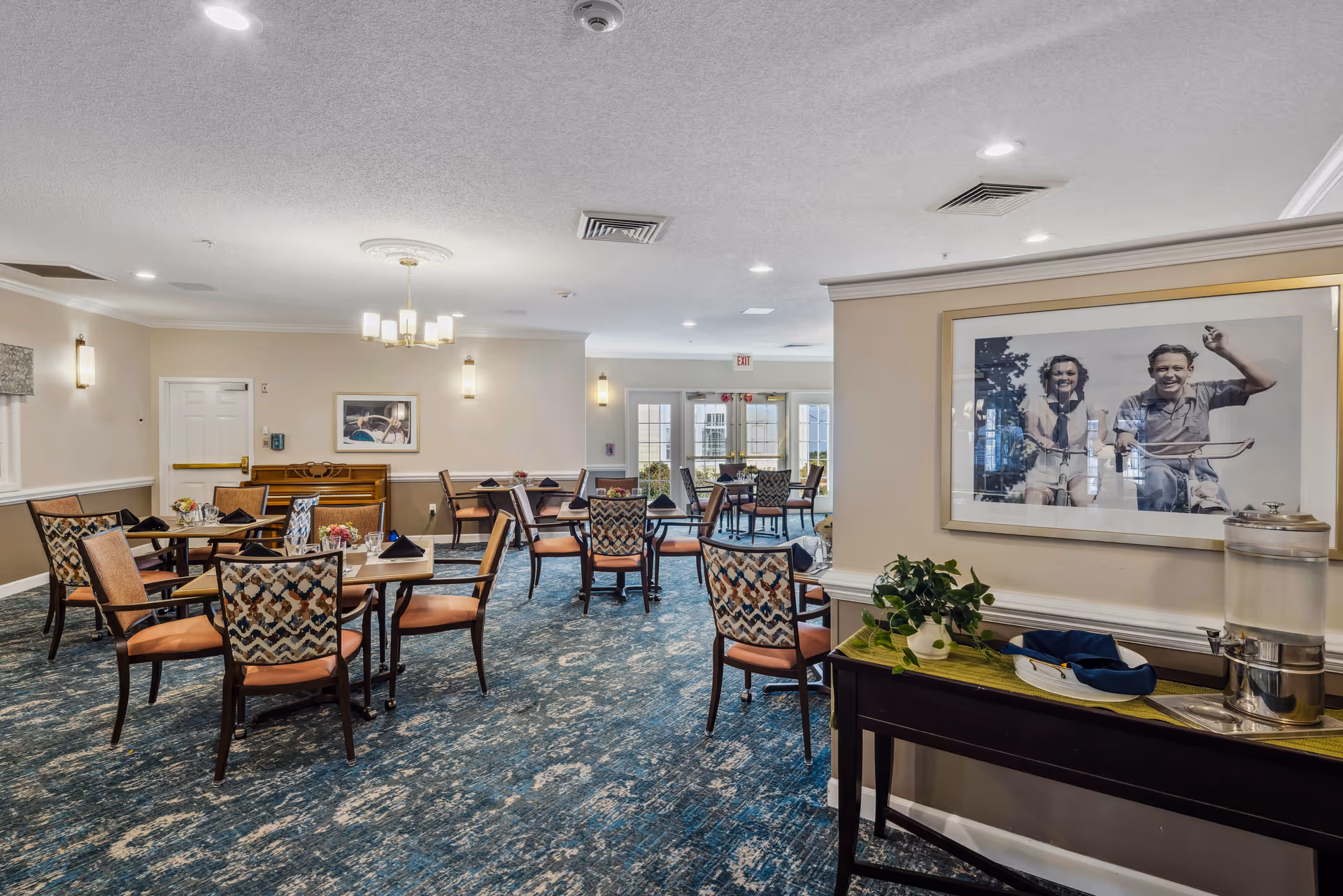 Spacious communal dining room with multiple tables and patterned chairs, a sideboard with a water dispenser, and framed artwork on the walls.