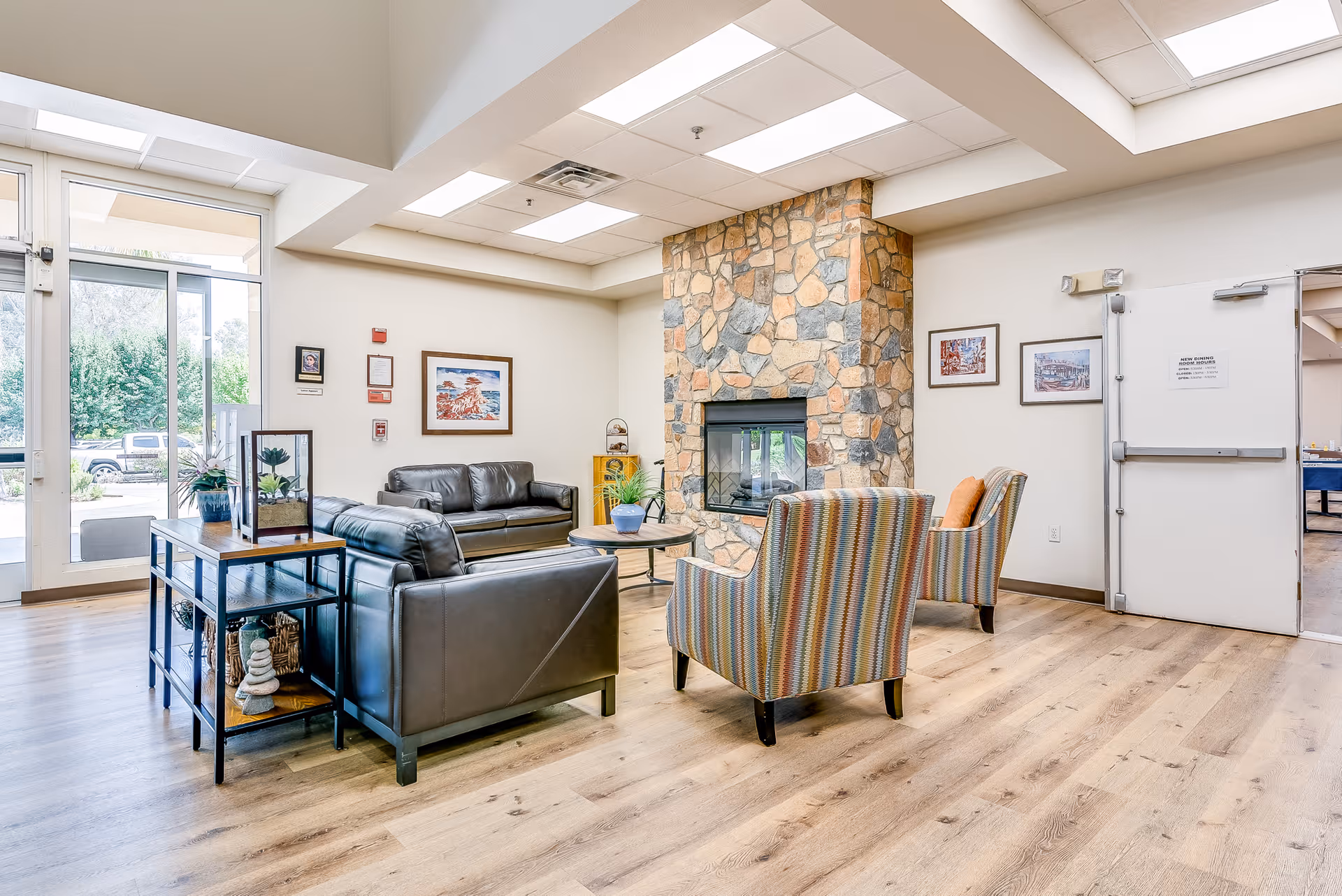 Bright senior living facility common lounge with sofas and armchairs arranged around a stone fireplace and large windows.