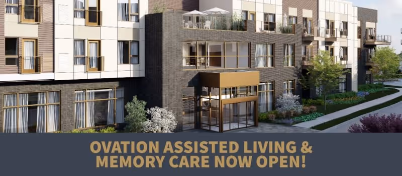 Exterior view of a modern multi-story assisted living facility with large windows, balconies, and landscaped greenery along a sidewalk.