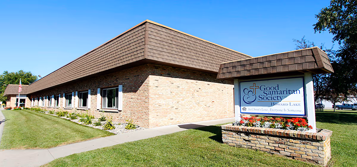Exterior view of the Good Samaritan Society - Howard Lake building, a single-story brick structure with multiple windows and a shingled roof. In front of the building is a sign with the facility's name and slogan, surrounded by a flower bed. The sky is clear and blue, and there is a well-maintained lawn and sidewalk leading to the entrance.